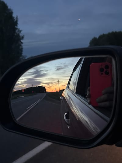 The image depicts a view through a car's side-view mirror at dusk. The mirror reflects a road stretching into the distance, lined with trees and illuminated by the warm hues of a sunset. Traffic signs are visible along the roadside, and power lines traverse the landscape. A small, pale moon is visible in the darkening sky. Within the car, a person's hand holds a bright red smartphone, also reflected in the mirror. The car's interior, including a door handle, is also visible. The scene appears to be set in or near the city of Yekaterinovka, Russia.