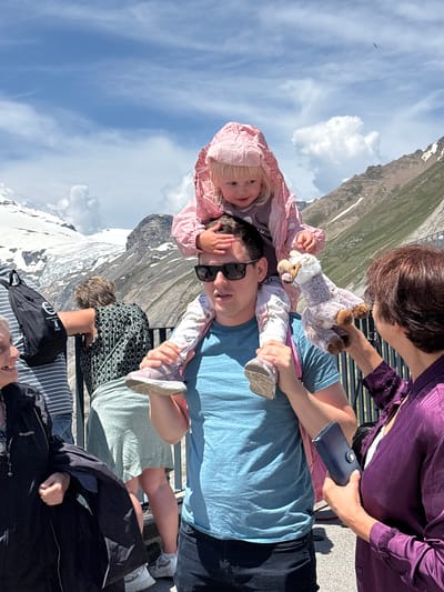 A medium shot captures a group of people outdoors with a mountainous, snow-capped backdrop. In the foreground, a man with sunglasses carries a young child on his shoulders. The child, wearing a pink hooded jacket, holds a stuffed animal. A woman to the right, wearing a purple top, is interacting with the child. Other individuals are visible in the background, and a black backpack is slung over one person's shoulder. The environment suggests a tourist location in Heiligenblut am Großglockner, Austria, given the presence of glaciers and mountains.