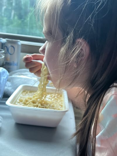 A close-up shot shows a young person eating ramen noodles from a white styrofoam container. The person's face is turned to the side, and their hair is partially obscuring their features. The ramen noodles are being lifted from the container with a white fork. In the background, a can of soda and a window with a blurry outdoor scene are visible. The scene appears to be indoors, possibly on a train or in a dining car, given the window view and the disposable packaging of the food. The location is not identifiable as Kizner, Russia.
