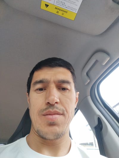 This is a medium shot, selfie-style photo of a man in a car. He is looking directly at the camera with a neutral expression. The man has dark hair, dark eyebrows, and a light stubble beard. He is wearing a white t-shirt. The interior of the car is visible, including the grey headliner, a grab handle, and a section of the car door with a black seatbelt. A yellow sticker with black text and a warning triangle symbol is attached to the car's headliner. The text on the sticker is in both English and what appears to be Chinese or Japanese characters. The lighting suggests it is daytime, and the background visible through the car window is blurred, but appears to be an outdoor scene.
