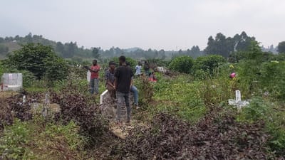 A cloudy day over a graveyard in a field. Several people are present amidst the overgrown grass and headstones. Some stand or sit on the graves, chatting. The graves are simple, mostly white, amidst dense vegetation. Trees cover the distant hills, with overcast sky above.