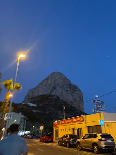 The image depicts a street scene in Calp, Spain, at dusk or early evening. A large, distinctive rock formation, known as Ifach Rock, dominates the background. In the foreground, a streetlamp with a bright yellow light illuminates the scene. To the right, a yellow building houses a restaurant named "Arroz Mariscos y Pescados," indicated by signage. A blue sign above the restaurant reads "Puerto Ifach Restaurante." Several cars are parked along the street, including a silver SUV and a dark-colored hatchback. A person, seen from behind, is walking away from the camera on the left side of the street, wearing a light blue shirt. The sky is a deep blue, suggesting twilight. Palm trees are visible on the left side of the image.