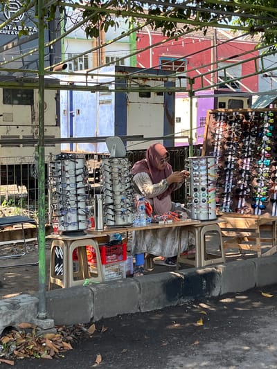 The image captures an outdoor market scene in Sumokali, Indonesia, focusing on a vendor selling eyewear. A woman wearing a pink hijab and a patterned dress stands behind a stall laden with various glasses. In front of her, a rotating display rack holds numerous pairs of eyeglasses. To her right, a larger display board showcases a wide assortment of sunglasses. A table in front of her holds a few more pairs of glasses, a bottle of water, and other miscellaneous items. The stall is set up under a canopy of trees, with a backdrop of buildings exhibiting a mix of colors including red, purple, and white, hinting at a busy urban environment. The ground is paved and slightly uneven, with fallen leaves scattered near the curb.