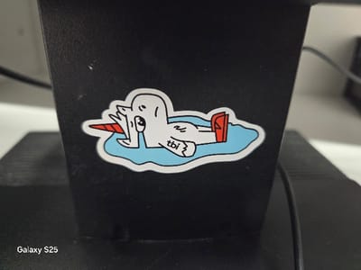 A rectangular sticker is affixed to a matte black vertical surface. The sticker depicts a cartoon illustration of a white, duck-like creature with a red conical horn on its head, lying on its back in an irregular light blue shape resembling a puddle. One of the creature's webbed feet is adorned with a red boot. The letters "tbi" are printed on its torso. The matte black vertical surface exhibits minor white specks and scratches. Below the sticker, the white text "Galaxy S25" is visible on a horizontal black surface. The background is out of focus, revealing blurred elements consistent with an indoor setting, including a dark object and a black cable to the right, and a lighter, indistinct surface to the left. No individuals or dynamic actions are depicted. The image does not provide visual cues to determine a specific location within Novo Delchevo, Bulgaria.