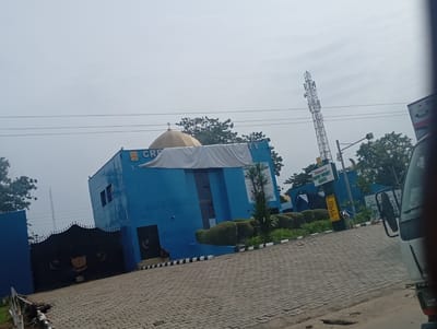 A blue building with a golden dome and the word "CREA TY" partially visible is observed. A white cloth is draped over the building's front. To the left, there is a dark gate with crescent moon and star symbols. A cell tower stands in the background. Paved ground covers the foreground. A sign reading "Welcome to Crescent University Abeokuta" is visible on the right side, indicating the location is in Abeokuta, Nigeria. A vehicle is visible in the foreground on the right. The sky is overcast.