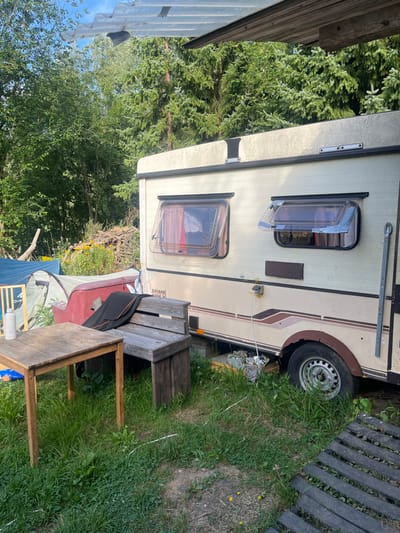The image captures an outdoor scene in Sint-Pieters-Leeuw, Belgium, depicting a semi-permanent encampment. A beige and brown Esterel caravan is positioned centrally, exhibiting two clear acrylic windows on its visible side, both propped open; the left window shows internal red and white fabric, and the right reveals darker material. The caravan's exterior is discolored and shows wear, with a visible rusted wheel well above a single silver-rimmed wheel on the lower right. A white electrical cable extends from an external outlet on the caravan's side.
In the foreground, a light-brown wooden table is situated to the left, with a white water bottle on its surface. To the table's right, a weathered, dark-grey wooden bench holds a folded black garment with orange trim. The ground is covered in green grass and weeds. A corrugated translucent roof panel extends from the top of the frame, providing partial overhead cover, supported by a wooden beam on the right.
To the left of the caravan, a light-colored tent is pitched, and behind it, a reddish-brown object, possibly a boat or cover, is visible. A partial view of a wooden chair is present near the tent. The background consists of dense green foliage, primarily coniferous trees, suggesting a wooded or rural setting. A large pile of wooden debris is discernible behind the tent. No individuals are present in the frame.