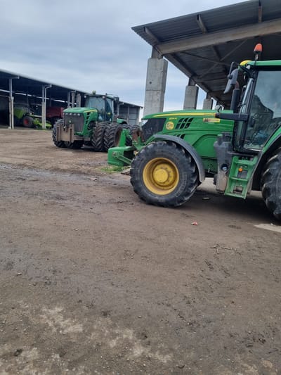 The image displays a row of large agricultural tractors parked under a substantial open-sided shelter in Ovcharovo, Bulgaria. The main focus is on a green John Deere tractor in the foreground, easily identifiable by its prominent green body with yellow detailing on the wheel hubs. Visible text on this tractor includes "JOHN DEERE," the model number "6155M," and "МЕГАТРОН" (MEGATRON), along with a "5" logo. The large front tire shows "MICHELIN" and the size "560/65R-28."
Further along the row, several other green tractors are parked, including one with a visible red wheel, suggesting it might be a CLAAS model. The shelter under which the machinery is stored features a corrugated metal roof supported by sturdy concrete pillars.
The ground beneath and around the tractors is a dirt and gravel surface, appearing damp or muddy in areas. The sky is overcast, indicating a cloudy day. No people are present, and the tractors appear to be idle or stored, awaiting their next use or maintenance. The scene depicts a typical agricultural storage or service area for farm equipment.