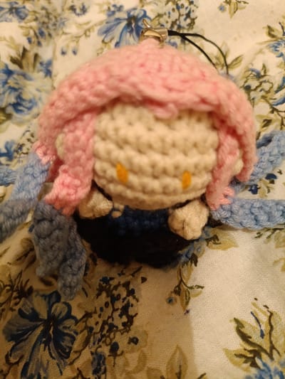 The media file shows a close-up, overhead view of a crochet amigurumi plush toy. The toy is predominantly cream-colored with a pink crocheted headpiece that resembles hair. Two small, orange dots on the face serve as eyes. Blue crocheted appendages, likely tentacles or limbs, extend outwards from the sides. The bottom of the toy appears to be a dark blue or black crocheted base. A small metal loop and a black string are visible at the top, suggesting it might be a keychain or ornament. The toy is placed on a fabric with a floral pattern featuring blue and white flowers. The lighting is dim, and the focus is on the toy, with the background slightly blurred. No text is visible. The setting appears to be indoors, on a piece of fabric. The overall mood is cute and whimsical due to the nature of the amigurumi.