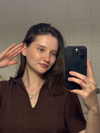 The image is a vertical, selfie-style photograph taken indoors. The primary subject is a young woman with long, dark brown hair, fair skin, and light-colored eyes. She is wearing a dark brown, textured knitted top and a delicate silver necklace with a cross pendant. Her right hand is holding a black smartphone to take the picture, and her left hand is touching her ear.
The background appears to be a tiled wall, suggesting a bathroom or a similar interior space. The lighting is soft and somewhat dim, possibly indicating evening or a poorly lit room. The woman's expression is neutral, with a slight tilt of her head. There is no visible text in the image. The overall impression is of a casual, personal snapshot. The provided location context of Журавлево (кемеровский), Russia, places the setting in a specific region of Russia, though there are no direct visual cues within the image to confirm this location.