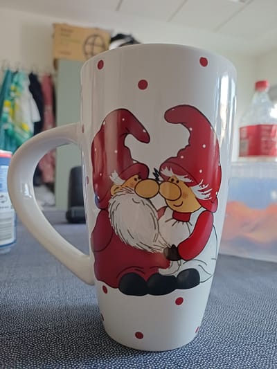 A close-up, eye-level shot shows a white ceramic mug with red polka dots and two cartoon gnomes kissing. The gnomes wear red pointed hats with white polka dots, red jackets, and white beards. The mug's handle is on the left. The mug sits on a textured blue surface, likely a tablecloth. In the blurred background, clothing hangs on a rack, a cardboard box with a circular logo is visible, and a plastic bottle with a red cap is on the right. The setting appears to be a domestic indoor environment. This image is not specific to Ballerup, Denmark, as no identifying landmarks or contextual elements suggest that location.