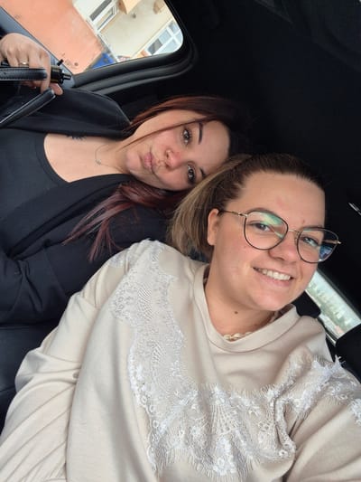 Two women are in a car. The woman on the left has dark hair with red highlights, a nose ring, is wearing a black top and jacket, and is pursing her lips. She is holding the steering wheel. The woman on the right has blonde hair pulled back in a ponytail, wears glasses, is smiling, and is wearing a tan sweatshirt with white lace. The background shows a glimpse of the outside world, including buildings, with one appearing to be orange. The window offers a view of what appears to be buildings in Villerupt, France.