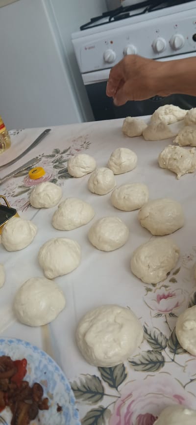 The image shows a close-up view of several small, round dough balls arranged on a table covered with a floral-patterned tablecloth. A person's hand, with fingers slightly wet, is visible in the upper right corner, suggesting the dough is being prepared or shaped. In the background, a white stove with burners and knobs is partially visible, indicating a kitchen setting. The overall scene is consistent with food preparation in a domestic environment. The location in the city of Mihaylovka, Kyrgyzstan is not discernible from the image itself.