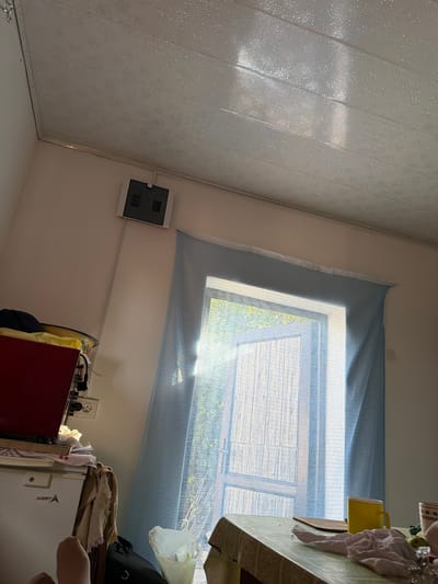 This image depicts an interior scene, likely a room in Vostok, Kyrgyzstan. A table covered with a green patterned tablecloth is in the foreground, with a yellow mug, some crumpled fabric, and a wooden cutting board on its surface. To the left, partially visible, is a white refrigerator with the brand "Aventa" on it, and to its right, a red appliance. Above the refrigerator, a dark-colored bag is visible. Further to the left, a gray electrical box is mounted on the wall. A light blue sheer curtain hangs across a window that shows greenery outside, indicating natural light is entering the room. The ceiling is a white, patterned paneling. The overall impression is of a lived-in domestic space.