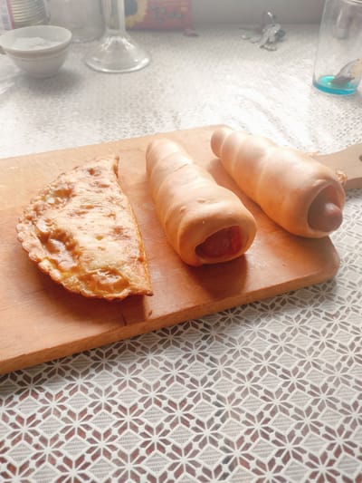 On a wooden cutting board, placed on a white lace tablecloth, are three savory pastries. To the left is a golden-brown, half-moon shaped pastry, likely fried, with a crimped edge. To the right are two pastries wrapped in golden-brown dough, resembling horns. Both are open at one end, revealing a sausage inside each. The pastry on the left is closer to the viewer, and the two sausage-filled pastries are positioned behind it and to its right. The background is blurred, showing what appears to be kitchen items and a window. This image was taken in a kitchen in Salsk, Russia.