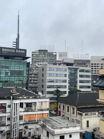 The image depicts a dense urban landscape in Abidjan, Ivory Coast, characterized by multiple buildings of varying architectural styles and heights under an overcast sky. The foreground features the dark, corrugated roof of a building with satellite dishes and a chimney. Adjacent to it is a white, multi-story building with balconies, striped awnings, and visible air conditioning units. Further back, a modern, glass-fronted building with the "BRIDGE BANK GROUP" sign is prominent. To its right, a taller, grey building with many windows stands, with a smaller sign displaying "AITEK" visible. In the background, more complex and older buildings can be seen, some with visible scaffolding. Billboards with logos for "AFG BANK" and "ISIS LOUE" are also present. The scene suggests a commercial and residential area within a busy city, with a focus on architecture and infrastructure. There are no discernible people in the image, nor are any specific actions or interactions taking place. The overall impression is a slightly hazy, grey urban environment.