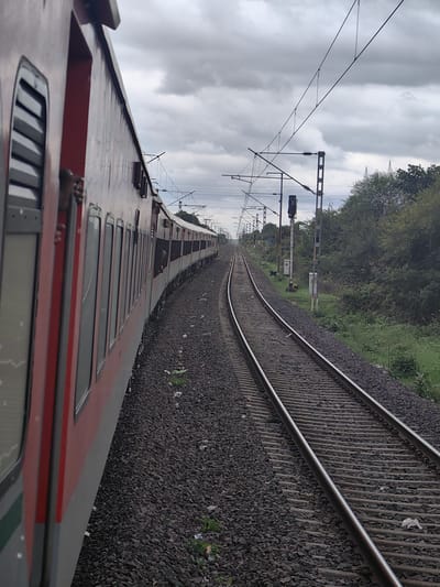 An eye-level, full shot captures a train in motion on a cloudy day in Khandwa, India. The perspective is from inside the train, looking out the side of a carriage. The train's red and grey exterior is visible on the left, with several windows. A person's hand can be seen resting on the windowsill of one of the open windows. The train tracks stretch forward and curve to the right, bordered by dark grey gravel. To the right of the tracks, lush green trees and vegetation line the embankment. Overhead, a complex network of electrical wires and poles supports the train's power supply. A traffic signal with a black 'X' symbol is visible in the distance, along with several utility boxes. The sky is filled with heavy, grey clouds, suggesting overcast weather.