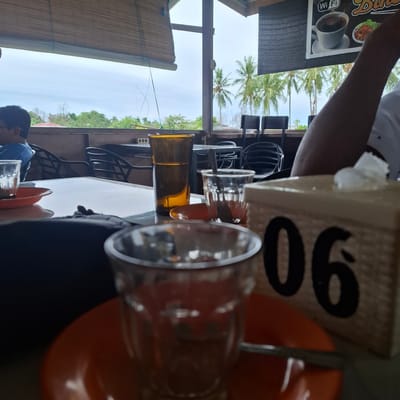 The image captures an outdoor dining scene at a restaurant in Mesjid Tuha, Indonesia. In the foreground, a reflective tabletop holds several objects, including an orange saucer with a small glass, a clear glass with a spoon, and a tall amber glass with a liquid. To the right, a tissue box with the number "06" printed on it is partially visible. A person's arm, dressed in a white shirt, extends into the frame from the right. In the background, several empty black chairs are arranged around tables, and beyond them, a view of palm trees and a cloudy sky is visible through an open-air structure. A person wearing a blue shirt is seated at a table on the left. The restaurant appears to be situated with a view of natural scenery.
