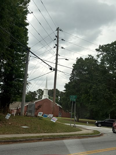 The image captures a street scene, likely in a residential area, beneath an overcast sky. Power lines crisscross the view, suspended between utility poles. The foliage is abundant, with mature trees framing the sky. A church is visible in the background, identifiable by its steeple. Several political campaign signs are planted in the grassy area adjacent to the sidewalk. Street signage indicates the location is at the intersection of Cole Drive and Five Forks Trickum Road. The OCR identifies that the image is near the city of Mountain Park, Georgia, USA. A vehicle is partially visible on the street.