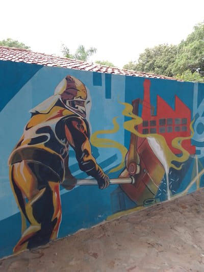 The image depicts a mural on a blue wall in Villa Hayes, Paraguay. The mural features a stylized illustration of a worker wearing protective gear, seemingly manipulating a large ladle filled with molten metal. Yellow fumes rise from the ladle. Behind the ladle, a red industrial building with multiple windows is depicted. The background is a light blue color. The wall is topped with a red tile roof, and foliage is visible above the roofline. The base of the wall rests on a concrete surface.