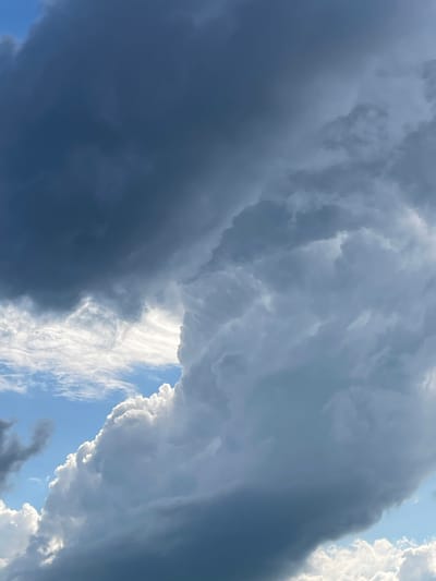 The image shows a dramatic sky filled with dark, ominous storm clouds. The clouds are predominantly gray and blue, with some areas appearing almost black. There are patches of bright white clouds in the foreground, illuminated by sunlight, creating a stark contrast with the darker clouds. A small sliver of blue sky is visible between the cloud formations. The composition is abstract, focusing solely on the textures and dramatic lighting of the clouds. There are no discernible people, objects, or structures in the image, nor any indication of human activity or specific location within the city of Маминское, Russia. The scene depicts a natural weather phenomenon.