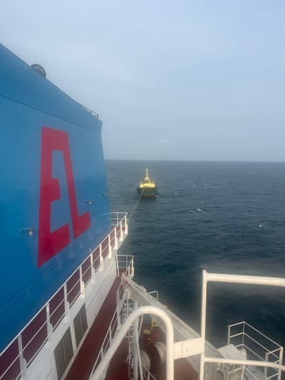 A tugboat is towing a massive ship. The ship has a blue exterior with a large red symbol. The ocean stretches out, a dark blue expanse beneath a light blue sky. The photograph is taken from an elevated position on the ship, capturing the deck's railing and machinery in the foreground.