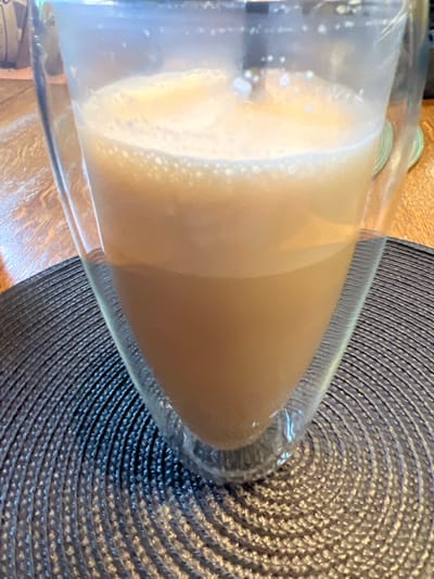 A close-up, vertical shot shows a double-walled glass mug filled with a frothy, light brown liquid. The mug is positioned on a dark, circular placemat with a textured, woven pattern. The placemat sits on a wooden surface. The liquid inside the mug appears to be a beverage like coffee or tea with milk, as evidenced by the foam and light brown color. The top portion of the liquid is foamy and bubbly, while the lower portion is smoother and darker. The glass mug has a distinctive conical shape, wider at the top and tapering towards the bottom. The lighting suggests an indoor setting, likely during the daytime, with some ambient light creating reflections on the glass. There are no people or animals visible in the image, and no discernible landmarks or environmental clues that would indicate a specific location within Berehove, Ukraine.