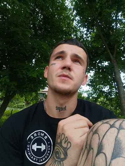 This is a medium shot, low-angle selfie of a young man with short, dark hair, looking upwards and to his right. He has a tattoo on his neck, which appears to read "soul" in gothic font, and another tattoo on his left cheek, which is partially obscured. He is wearing a black t-shirt with a circular logo on the left chest. The logo is white and contains an image of a dumbbell in the center, surrounded by text that reads "BE MORE. TRAINING. PERSONAL." in a circular pattern. He has extensive tattoos on his left arm, including an eye with radiating lines and a spiderweb design on his leg.
The man is outdoors, with lush green trees and foliage in the background. The lighting suggests it is daytime, possibly late afternoon, with dappled sunlight filtering through the leaves. The overall mood appears relaxed and contemplative, with the subject gazing upwards. The setting appears to be a backyard or a park, with a hint of a fence and some vegetation in the distance, suggesting a rural or suburban environment. The image context places the location as Soroca, Moldova.