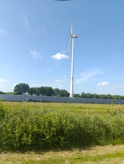 In Heidenau, Germany, a field of solar panels stretches across the landscape, bordered by a line of trees. To the right of the solar farm, a tall white wind turbine stands prominently against a bright blue sky with scattered white clouds. Further in the distance, several other wind turbines are visible. The foreground is a blur of green foliage, suggesting movement.