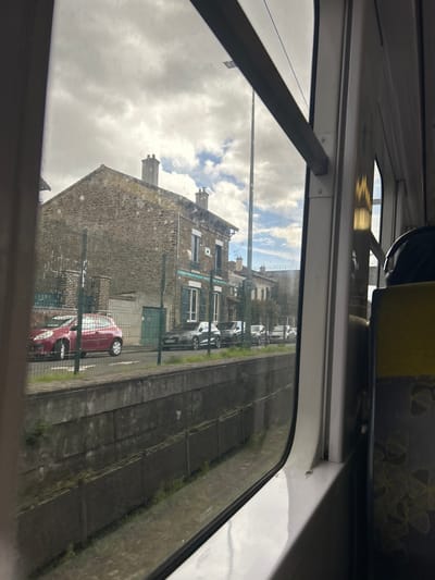 The image depicts an exterior urban scene in Arcueil, France, observed from inside a vehicle, likely a train or tram, through a window. The immediate foreground shows the white interior window frame and a portion of a yellow and dark grey patterned seat on the right. Reflections are visible on the window glass.
Outside the window, a concrete canal or trench with weathered grey walls, partially covered by sparse green vegetation, runs horizontally across the lower third of the frame. Above this, a green metal fence separates the canal area from a paved street. Several vehicles are parked along this street. Prominently, a red three-door hatchback is parked closest to the viewer, followed by a dark grey sedan or hatchback further down the street. Additional vehicles, lighter in color, are visible in the distance.
In the background, a row of multi-story buildings with stone facades, pitched roofs, and multiple chimneys lines the street. The most prominent building on the left features rough-hewn stone and a dark roof, while an adjacent building to its right has blue decorative trim around its windows and dark shutters. The sky overhead is partially overcast with large grey clouds interspersed with patches of blue sky and white cumulus clouds. A tall, slender utility pole extends vertically into the sky on the right side of the main building. No individuals are visible. The scene is static, captured from a moving perspective.