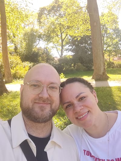 This outdoor selfie features two smiling individuals, a man and a woman, against a backdrop of bright green foliage and sunlight in what appears to be a park in Bayreuth, Germany.
The man, positioned on the left side of the frame (when viewed with the natural upright orientation), is bald with a light brown beard and mustache. He wears round, gold-rimmed glasses, which highlight his light-colored eyes, and is dressed in a white collared jacket over a dark top. He is smiling gently.
The woman, located on the right side of the frame, has dark hair pulled back. She is smiling broadly, revealing her teeth, and has a small hoop nose ring and a delicate necklace. She is wearing a white t-shirt with red text partially visible on the chest, which appears to spell out "YOU TRY TO".
The background consists of vibrant green grass, possibly with small yellow flowers, and several large trees with lush, bright green leaves. Strong sunlight filters through the tree canopy, creating a warm, bright glow and indicating a sunny day, likely during the morning or late afternoon. The setting suggests a relaxed and pleasant outdoor environment, characteristic of a park or garden. Both individuals appear happy and comfortable.
