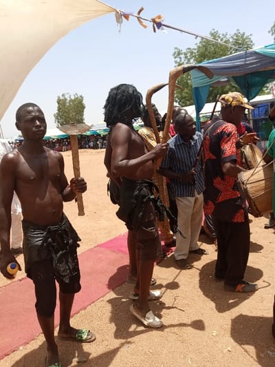 The image depicts a group of men in a public space in Zum, Nigeria, located at the coordinates 10.116 latitude and 13.046 longitude. The men are standing in a row, with the man at the forefront holding a wooden stick in his right hand. His left hand holds a blue container. The man is wearing a black loincloth and has no shirt on. The other men are all wearing clothing, some of which is traditional. One of the men wears a black loincloth and has his hair pulled back and tied. He appears to be holding a wooden stick. The ground is a light brown sand and there are red strips of cloth laid out on the ground. The environment appears to be a public gathering space with a few tents in the background.