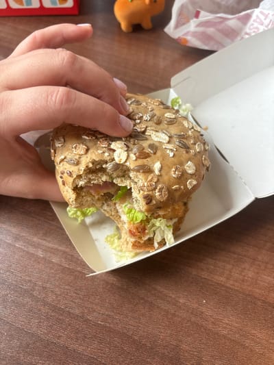 A high-angle, close-up indoor photograph depicts a person holding a partially consumed burger over a dark reddish-brown wooden table in the city of Krilovo, Russia. The burger, featuring a multi-grain bun topped with various seeds and oats, has a significant bite taken from its front. Visible fillings include shredded light green lettuce, a creamy white sauce, and layers of what appears to be pinkish meat and a yellowish component. The burger rests within a plain white cardboard container. A light-skinned hand with short, pale nail polish is positioned on the left side, grasping the bun. In the blurred background, to the upper right, a crumpled white paper bag with red stripes and partial text (e.g., "TAST") is visible. To the upper left, a small orange object, possibly a figurine, and a red object are discernible. The wooden table exhibits a distinct linear grain pattern.