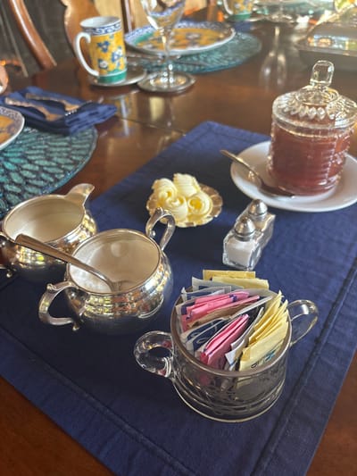 A meticulously arranged breakfast or dining table setting is depicted indoors, presumably in the city of Monterey, United States of America. The scene features a polished wooden table, partially covered by a dark blue fabric placemat or runner, and a teal-patterned circular placemat to the left.

In the foreground and midground, a collection of condiments and serving items is displayed on the blue placemat. On the far left, a silver creamer pitcher contains a white liquid. Adjacent to it, a silver sugar bowl holds white granulated sugar and a small silver spoon. To the right of these, a small scalloped silver dish presents three carefully shaped butter curls. A clear glass salt and pepper shaker set stands next to the butter dish. Further to the right, a clear glass jar with a decorative lid, containing a reddish-brown preserve (likely jam), rests on a small white saucer with a silver spoon. In the bottom right, a clear glass cup-shaped holder is filled with an assortment of individually wrapped sugar substitute packets in yellow, pink, and blue colors.

In the blurred background, parts of a place setting are visible, including a mug with yellow and blue floral patterns, a decorative plate with similar motifs, a clear wine glass, and silver cutlery (fork and knife) resting on a folded dark blue napkin. No individuals are present in the frame; the items are prepared for consumption.