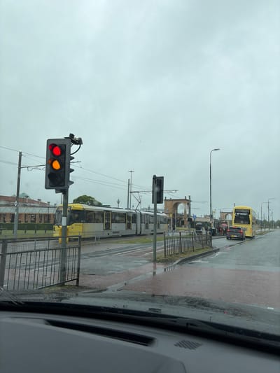 The image depicts a view from inside a vehicle looking out at a street scene in Trafford, United Kingdom, on an overcast day. A traffic light is prominently displayed in the foreground, showing red and amber lights illuminated. To the right of the traffic light, a yellow tram is stationary, with the number "307" visible on its side. Further down the street, a yellow double-decker bus is stopped at a bus stop, with several pedestrians, some holding umbrellas, waiting. A dark-colored car is also stopped behind the bus. In the background, there are buildings with arched structures and more distant structures. The road surface appears wet, indicating recent rain. A metal fence runs along the left side of the road, separating the pavement from the tram tracks and roadway. The overall environment suggests a public transportation hub in an urban setting.