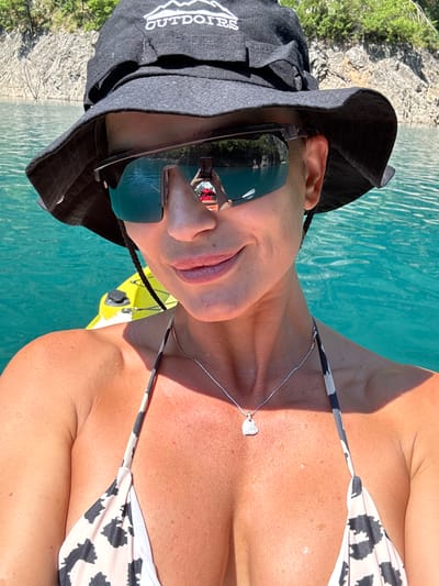 A woman wearing a black bucket hat with "OUTDOORS" embroidered on it, and a leopard-print bikini top, is kayaking on a bright turquoise body of water. She is wearing sunglasses that reflect a phone and a red object, and a silver necklace with a heart-shaped pendant. The background shows rocky, tree-covered hills. The water appears to be calm, with some ripples. The overall impression is one of leisure and outdoor recreation. This scene is likely in the area of Bezuje, Montenegro, known for its natural beauty and recreational water activities.