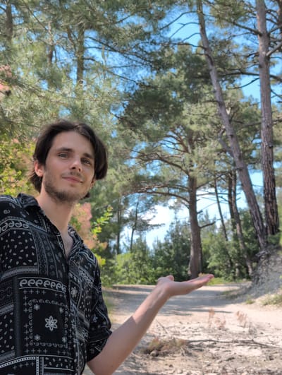 A young man with dark hair and blue eyes, wearing a black patterned short-sleeved shirt, stands in a wooded area. He is looking towards the camera with a slight smile and has his right hand extended, palm up, in front of him. The background consists of tall pine trees and a dirt path. The lighting suggests it is daytime. The location is identified as Dzhankhot, Russia.