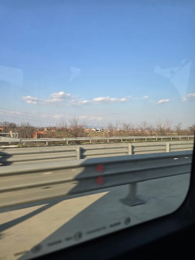 The image captures a daylight scene observed from a vehicle window, looking out over a highway or bridge in Opalchenets, Bulgaria. The upper frame is dominated by a clear blue sky, featuring a horizontal band of widely spaced, small white cumulus clouds at mid-level. The horizon line reveals a rural or semi-urban landscape with numerous bare deciduous trees and bushes, indicative of late winter or early spring. Several residential-style buildings with reddish-brown tiled roofs are visible amidst the vegetation. In the foreground, a grey concrete road surface is present, partially covered by shadows cast from the left. A prominent, two-tiered grey metallic crash barrier extends across the mid-ground, featuring two distinct red circular markings on the section closest to the viewer. A secondary, similar crash barrier runs parallel behind the first. The bottom right corner of the image shows the dark frame of the vehicle's window, with the window glass itself displaying faint reflections and unreadable white alphanumeric details. No individuals are visible.