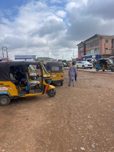 The image depicts a street scene in Ojodu, Nigeria. A row of yellow auto rickshaws, commonly known as "Keke Napep," is parked along a dirt road. One rickshaw is prominent in the foreground with its black canvas roof partly obscuring the driver. A person in patterned clothing is walking on the road. In the background, there is a building with signs for "CHRISVIC LAUNDRY," "MOZIRA GOLD," "GOD IS KING COMPLEX" and "BIG SHOW", indicating a commercial area. A white SUV is also parked nearby. A sign for "SPRINGFORTH SCHOOL" is visible attached to a lamppost or utility pole. Overhead, the sky is cloudy with patches of blue. Power lines are visible across the street.