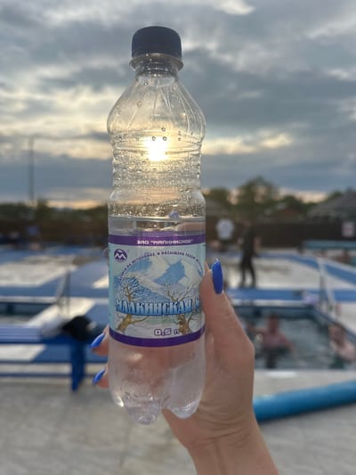 A person's hand with bright blue nail polish holds a clear plastic water bottle filled with water. The bottle has a label with blue and purple accents featuring a snowy landscape and text in Cyrillic. The sun is shining through the bottle, creating highlights. In the background, there is a swimming pool area with people in the water and lounge chairs. The sky is cloudy with the sun partially visible. The location is identified as Yuzhnyye Koryaki, Russia.