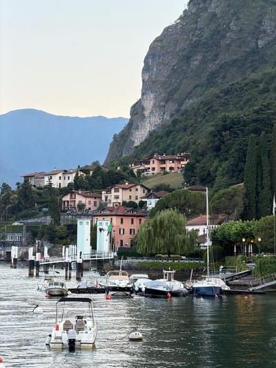 This is a photograph of the waterfront in Loveno, Italy. In the foreground, several boats are moored in the water. The boats are of various sizes and types, including a white motorboat with two seats in the back and several smaller fishing or leisure boats. Orange buoys are visible in the water. In the middle ground, there is a pier with white railings and a light blue structure. Several more boats are docked at the pier. Behind the pier, a row of buildings with terracotta roofs are situated on a hillside. The buildings are a mix of residential and commercial structures. A large, steep, rocky mountain covered in trees rises behind the buildings. The sky is a pale blue and appears to be either early morning or late evening. The overall scene is one of a tranquil lakeside town.
