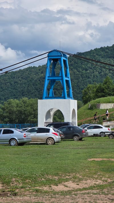 The image depicts a scene in the city of Tash-Asty, Russia, featuring a blue structure with white arched supports. The structure appears to be a pylon or tower, possibly related to a cable car system, with thick cables extending overhead. In the foreground, several cars are parked on a grassy area. Among the cars are a silver sedan, a silver hatchback, a dark gray sedan, and a white sedan with a trailer attached. Two individuals are walking up a set of stairs on the right side of the image, ascending a grassy embankment. The background consists of a densely forested hillside under a cloudy sky.