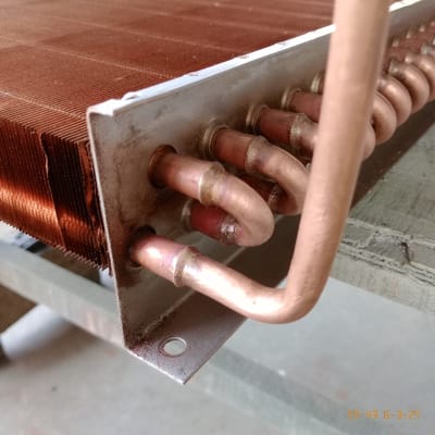 The image shows a close-up of a copper pipe system, possibly part of an air conditioning unit. The pipes are bent and connected to a metal plate with several holes. The copper pipes are covered in a layer of dirt and grime. The metal plate is also dirty and scratched. The location is Kecamatan Curug, Indonesia, based on the provided coordinates. The image was taken on a date that reads "05-43 6-9-25." There are no people in the image. The scene is a close-up of a section of the air conditioner. The image appears to be a snapshot of the system taken during a maintenance or repair process. It is unclear what actions are being taken.