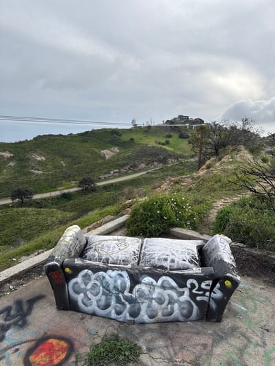 A graffiti-covered sofa sits precariously on a ledge overlooking a green hillside. A narrow road winds through the landscape, leading to a distant house. The sky is overcast with a mix of gray and white clouds. The concrete around the sofa is also covered in graffiti. Grass and small plants grow near the sofa.