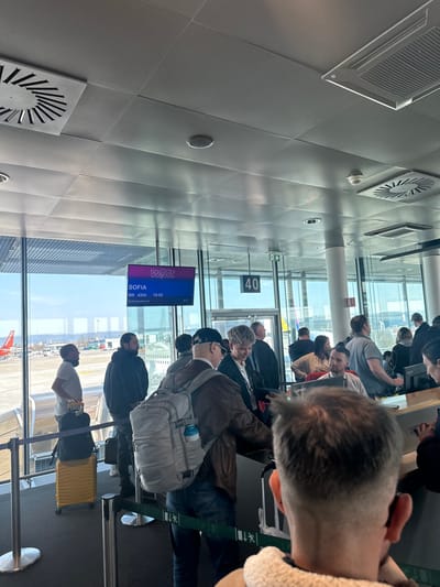 This image captures an indoor scene at Gate 40 of an airport, identified as EuroAirport Basel Mulhouse Freiburg in Saint-Louis, France, due to the combination of Wizz Air operations to Sofia and the specified location.

The environment features a modern aesthetic with a light-colored, segmented ceiling containing multiple circular ventilation grilles, recessed light fixtures, a square air conditioning unit, and a visible smoke detector. Large, clear windows extend across the background and left side, revealing an airport tarmac under a bright, clear sky. An orange-colored aircraft, displaying a red tail consistent with an EasyJet livery, is visible at a distant gate position. An electronic information screen centrally displays the "WIZZ" logo, "SOFIA" as the destination, flight number "W6 4324", scheduled time "15:25", and an ambient temperature of "4°C".

In the immediate foreground, the back of a male's head is visible, showing a short, faded haircut with darker hair on top and lighter sides. He wears a light-colored, possibly sherpa-lined, garment on his shoulders.

A queue is formed by approximately twelve to fifteen individuals, delineated by two parallel green and white striped retractable barriers. A prominent male passenger stands in the middle ground, wearing a dark brown leather jacket, blue jeans, and a dark baseball cap. He carries a large, light-gray tactical-style backpack with a blue water bottle attached. Beside him, a woman with short, light-colored, wavy hair wears a dark jacket over a light top, and appears to be smiling.

To the left, nearer the windows, a male with a dark beard and a black hoodie stands, looking towards the tarmac. Further left, another male in a light-colored t-shirt stands next to a bright yellow wheeled suitcase, also observing the tarmac.

On the right, a boarding desk is present, where an airport employee, partially visible behind a laptop with "Lenovo" branding, is likely processing passengers. Another male in a light grey t-shirt is visible behind the desk, looking towards the queue. Passengers are generally standing and waiting, indicating a boarding sequence for the identified Wizz Air flight to Sofia.
