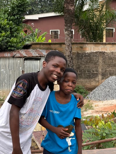 Two African children are standing outdoors in Orifite, Nigeria. The taller child wears a black and white shirt and smiles broadly, with a smaller child in a blue shirt wrapping an arm around his shoulder. Behind them is a mix of vegetation, including a palm tree, a corrugated metal fence, and concrete buildings. A pile of gravel or rocks is also visible.