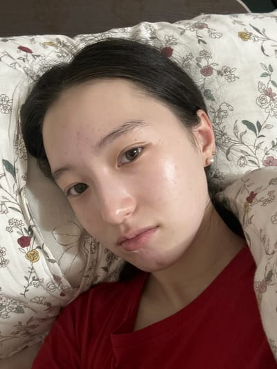 A young woman with dark hair and brown eyes lies on a floral-patterned pillow, her head resting on it. She is wearing a red shirt. Her skin is visible with some texture. The background appears to be a bed. The image is taken from a close-up perspective, focusing on her face and upper body. There is no indication of the city of Nookat, Kyrgyzstan in the image.