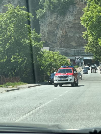 A red and white Toyota Hilux with flashing blue lights on its roof, identified by license plate KL CG071, is driving on a paved road in the city of Dulovine, Montenegro. The vehicle is traveling uphill alongside other cars. The road passes by a rocky cliff face covered in trees on the left and a tunnel entrance on the right. The scene is illuminated by daylight.