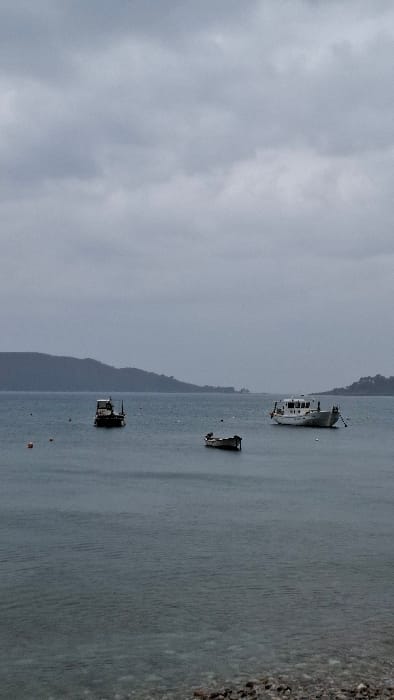 The image displays a coastal scene in Podbadac, Montenegro, under an overcast, uniformly gray sky. The water in the bay is calm, exhibiting a dark blue-gray hue with minor surface ripples. The immediate foreground shows a narrow strip of dark, wet pebbles forming the shoreline.
Three boats are present on the water. To the left, a dark-hulled, medium-sized utility boat with a lighter superstructure and cabin is anchored. To the right, a larger white vessel, possibly a fishing boat or small ferry, with a prominent forward cabin, is similarly moored. Centrally positioned between these two larger vessels, and slightly closer to the foreground, is a smaller, dark-colored, narrow rowboat or dinghy, appearing partially submerged. Several spherical buoys, colored orange and white, are scattered across the water's surface. The background features a distant, dark, undulating coastline covered with vegetation, extending horizontally across the frame. No individuals are visible, and no actions or interactions are depicted.