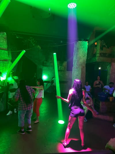 The image depicts an indoor scene at night, likely a nightclub or entertainment venue, characterized by vibrant green and purple lighting. In the foreground, a woman with dark, wet hair is dancing with her back to the camera, wearing a dark top and shorts. To her left, a woman in a plaid shirt and shorts dances, facing away from the viewer. A woman in red pants and a white top is to the left of the plaid-shirted dancer, facing towards her. Other individuals are visible in the background, some seated and some standing, amidst a setting that appears to have a jungle or cavernous theme, with stone-like pillars and textured walls. A "Grey Goose" bag is visible on a wooden structure in the background. The presence of a spotlight and numerous stage lights suggests a performance or active entertainment setting. While the specific location in Canet-en-Roussillon, France cannot be definitively determined from the image alone, the scene suggests a lively social gathering with music and dancing.