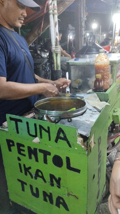 A person wearing a blue shirt and a grey cap stands behind a green food cart at night. The cart has "TUNA PENTOL IKAN TUNA" written on its side in large black letters. The person is holding a fork and appears to be serving food from a large metal pot. The cart is filled with various items, including bottles of sauce and what looks like money. In the background, other people and motorcycles are visible, suggesting a busy street or market setting. Lights from the food stall and surrounding area illuminate the scene. The time appears to be evening or night, and the weather is likely warm and humid, typical for Indonesia. The overall atmosphere seems lively, with the food cart likely operating in a local Indonesian market.